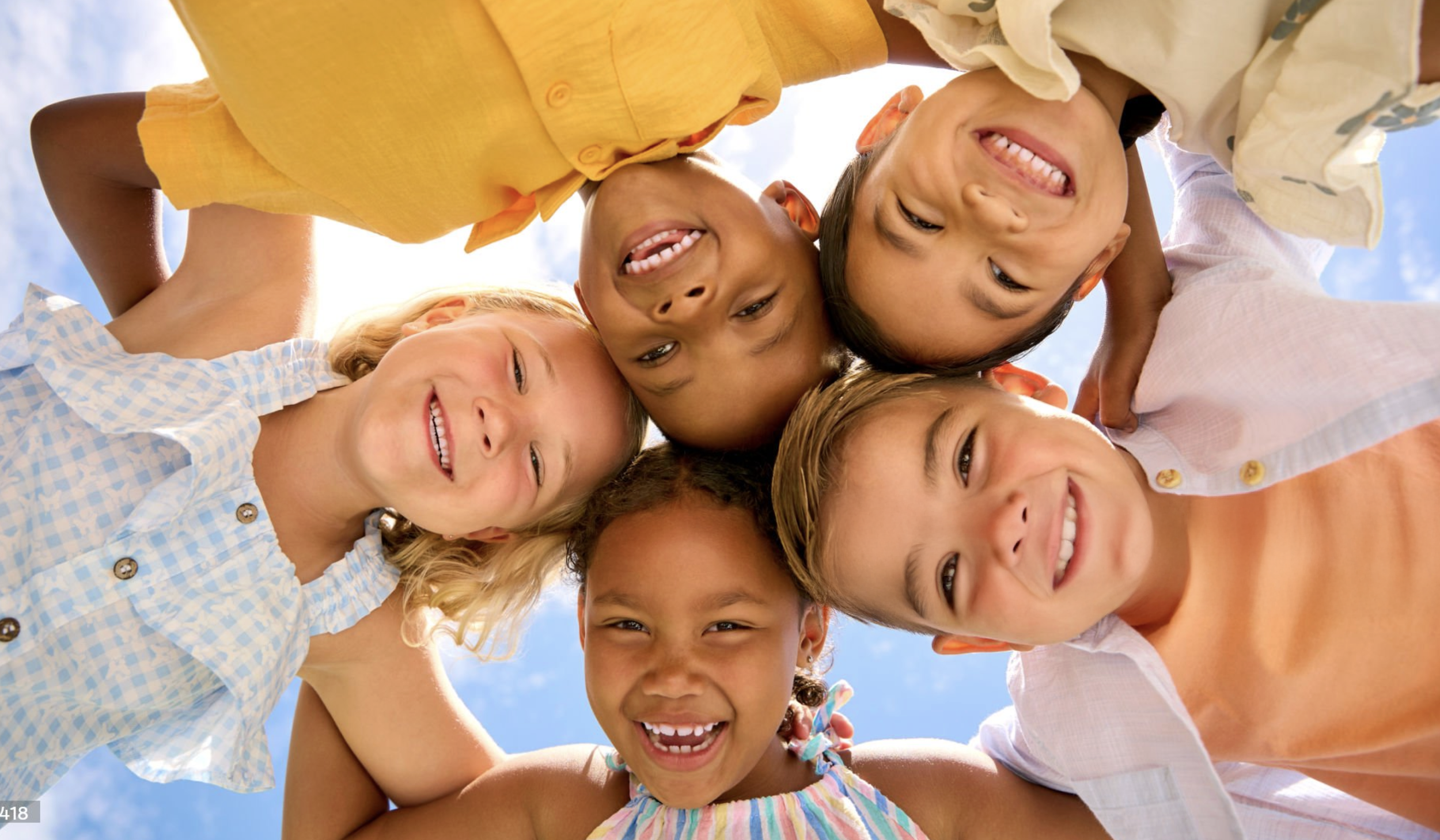 A group of six children looking down at the camera, smiling and laughing, with a bright blue sky in the background.