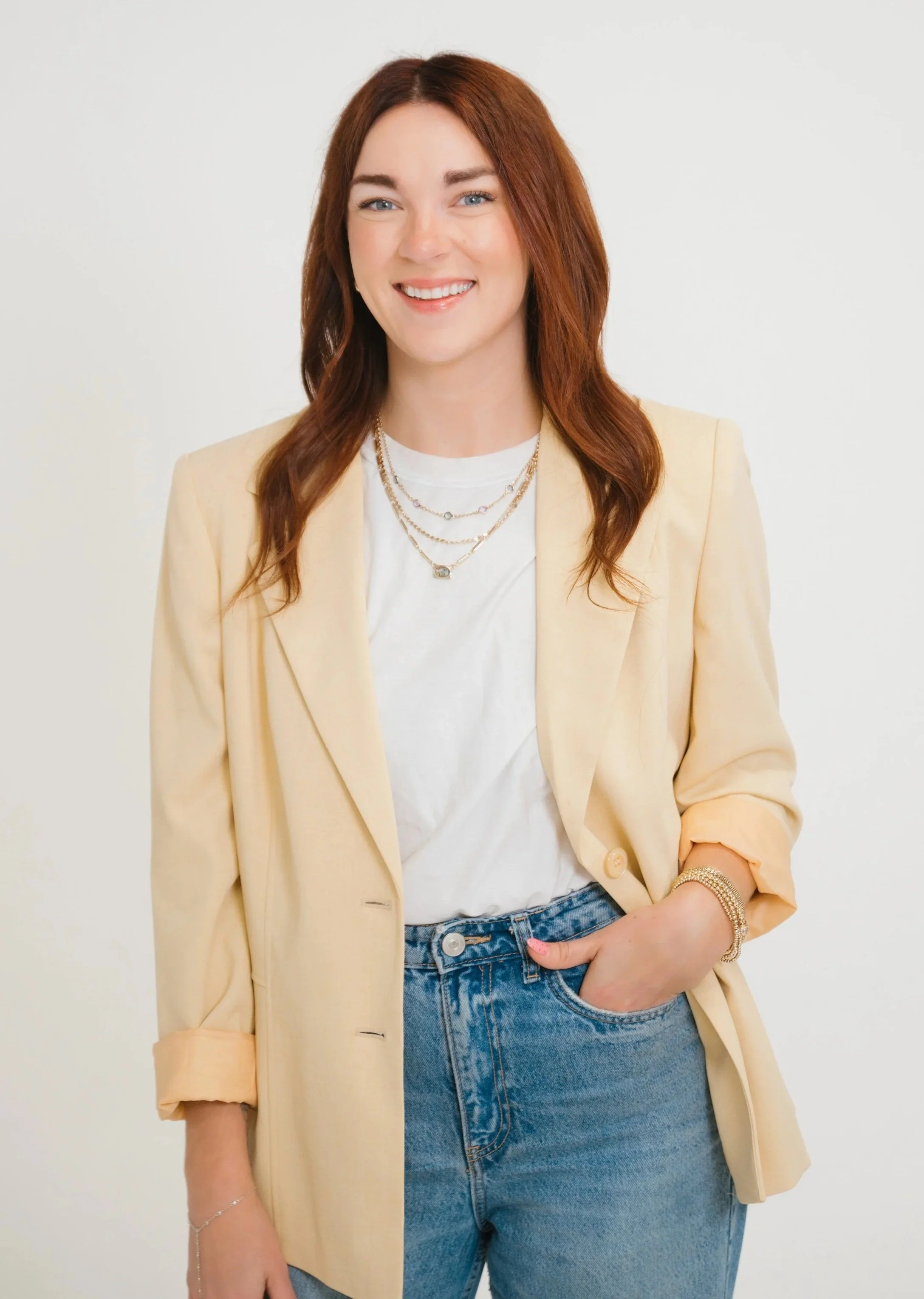 Board Certified Behavior Analyst Megan Alves  wearing a cream-colored blazer over a white t-shirt, layered necklaces, a bracelet, and blue jeans, standing against a plain light background.