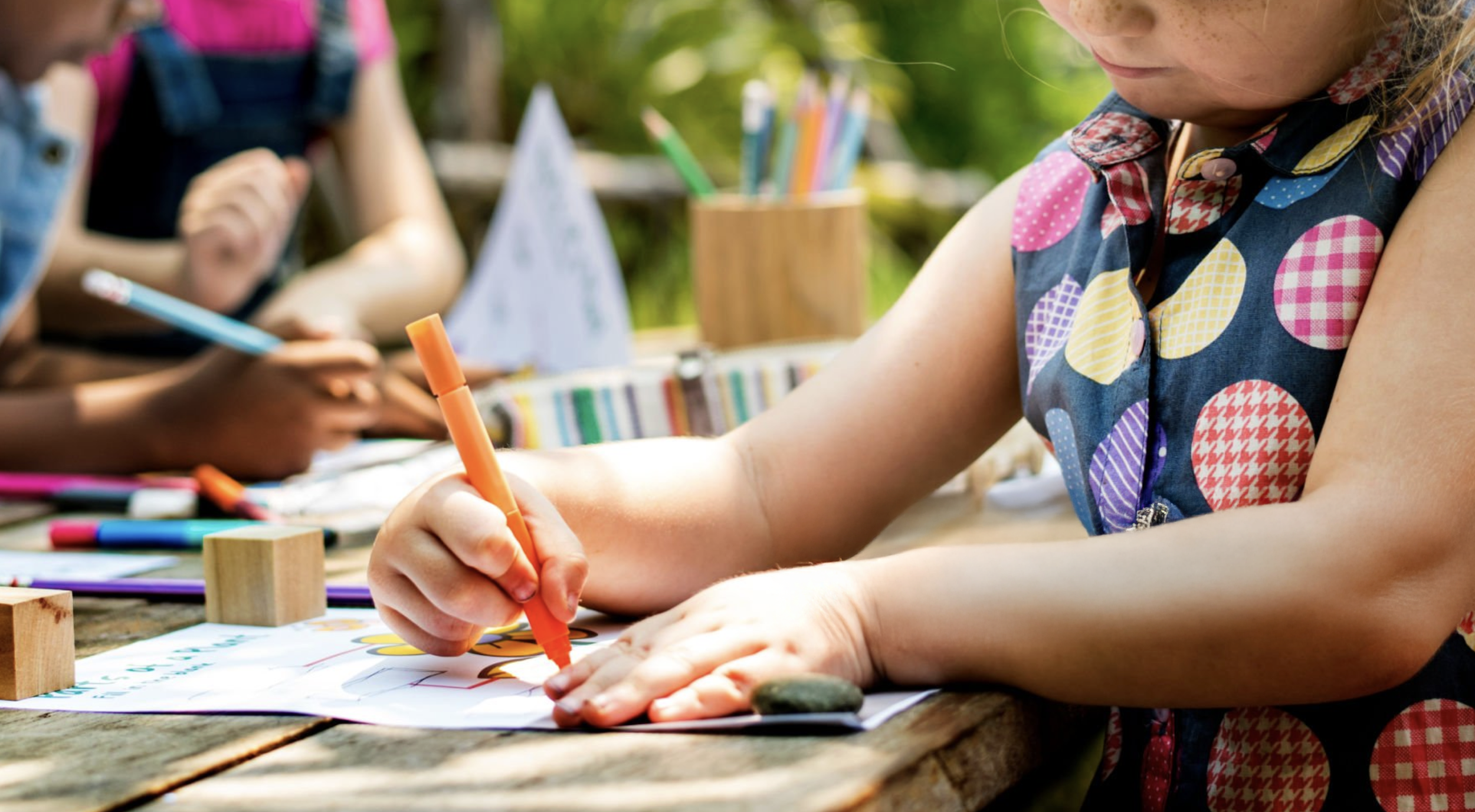 Child coloring with an orange marker at an outdoor wooden table, with children in the background also drawing and crafting.