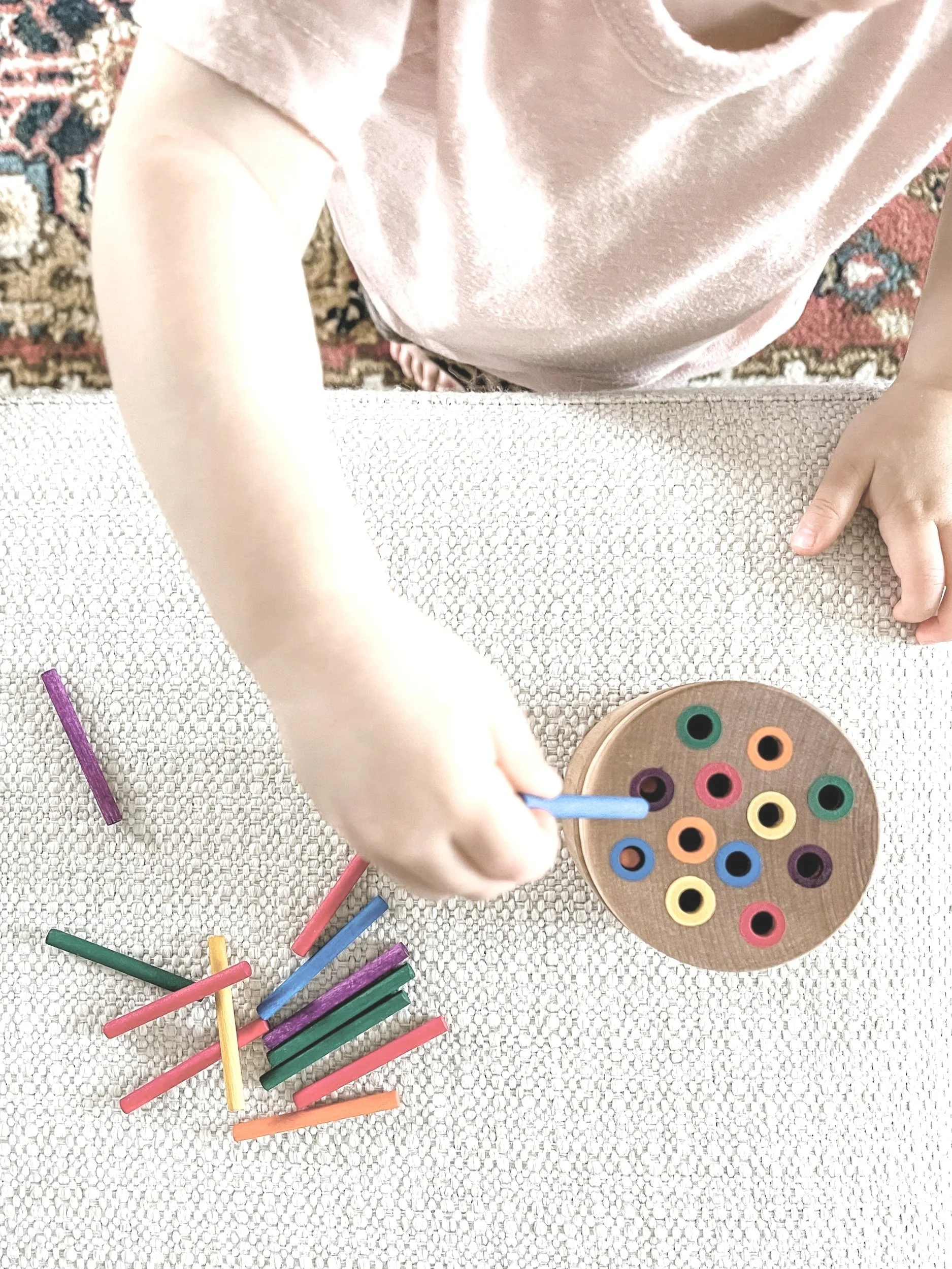 Child playing with colorful chalks and a wooden circle with holes filled with colored pegs on a textured white surface.