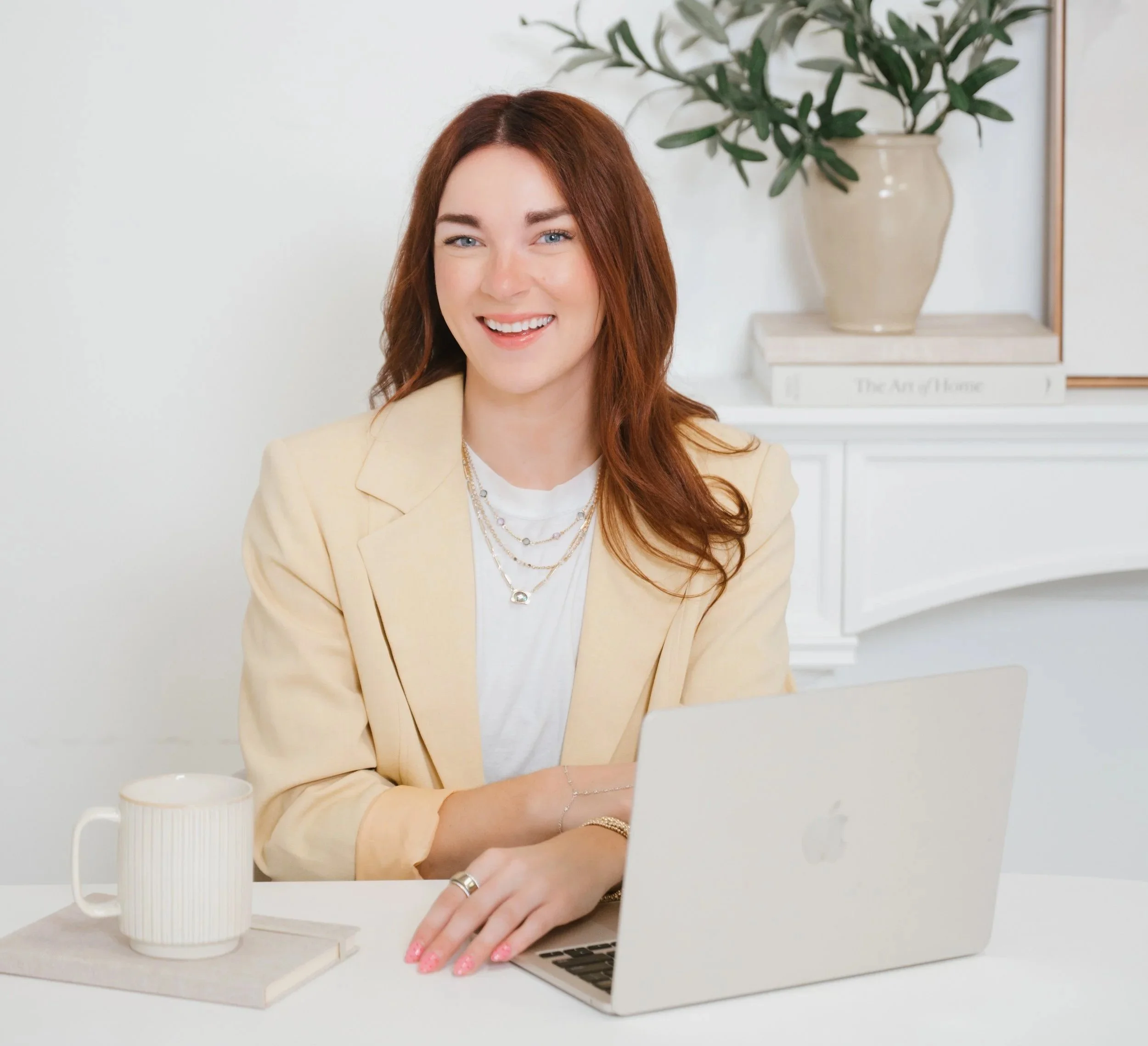 Board Certified Behavior Analyst Megan Alves sitting at a desk with an open MacBook computer, a white mug on a book, and wearing a yellow blazer and layered necklaces.