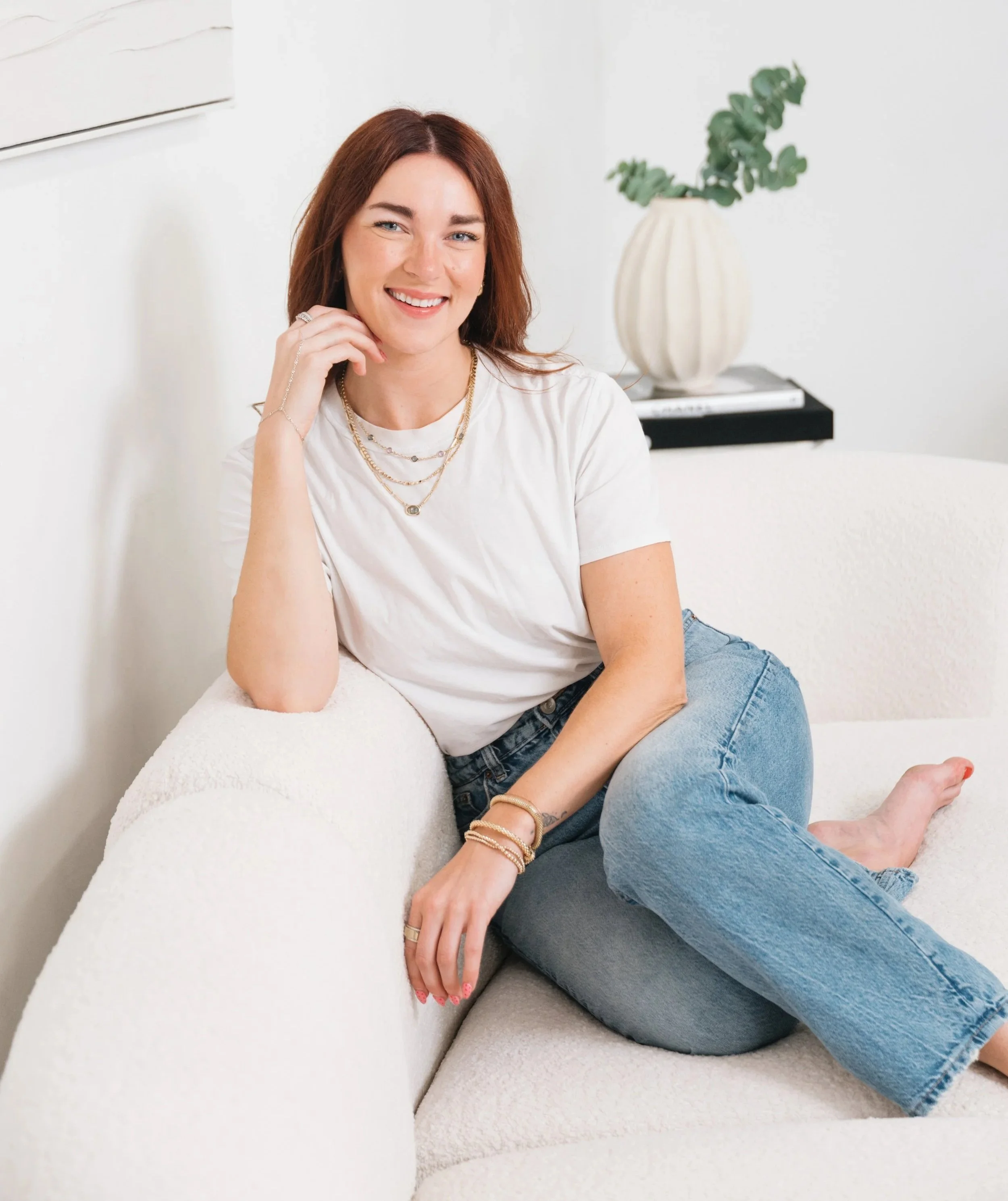 Board Certified Behavior Analyst Megan Alves sitting on a white couch, smiling, wearing a white t-shirt and blue jeans, with jewelry including necklaces and bracelets.