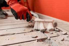Person wearing red gloves using a brush to break apart ceramic tile on a wooden floor.