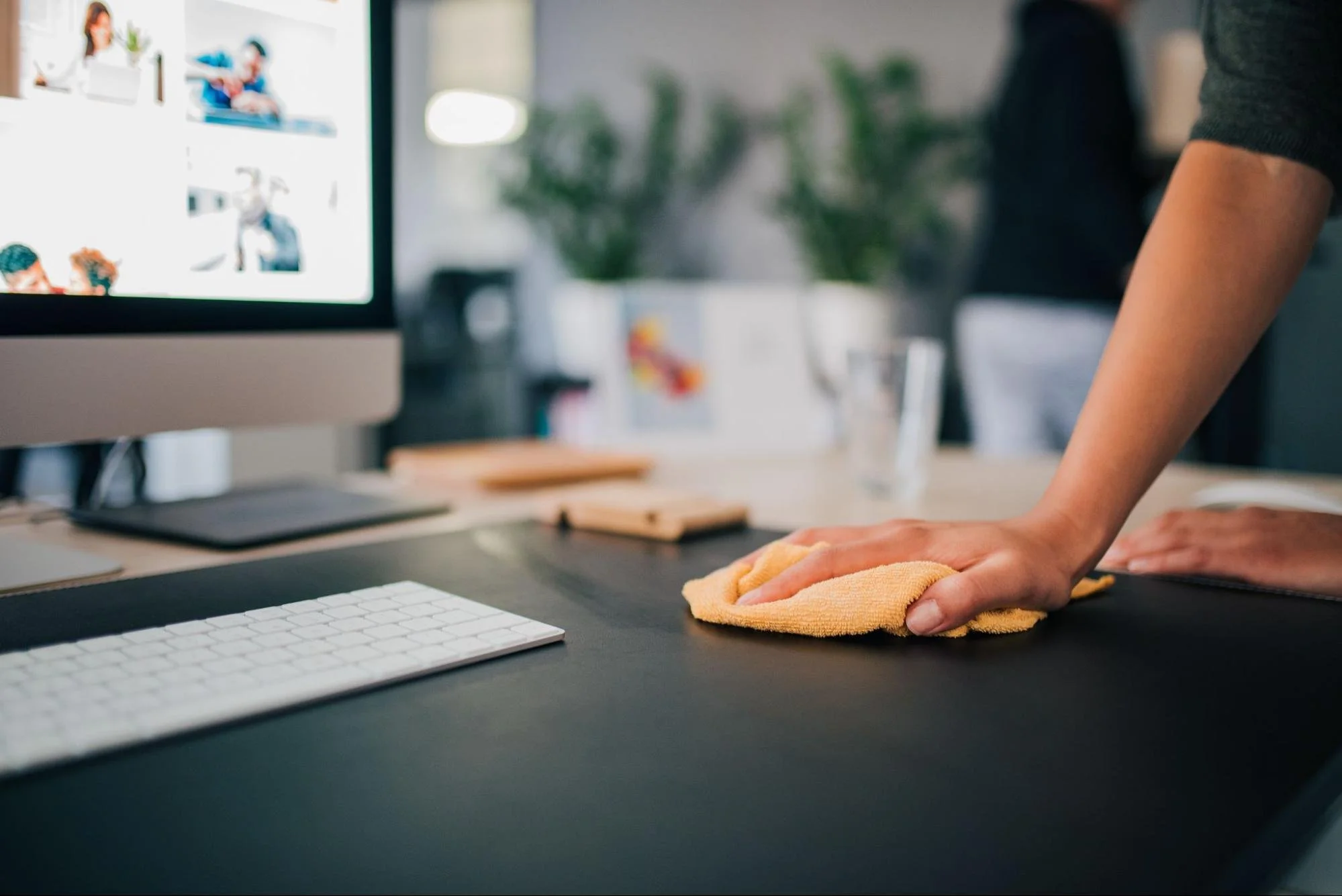 Person cleaning a computer desk with a yellow cloth, positioned next to a monitor, keyboard, and other office items.