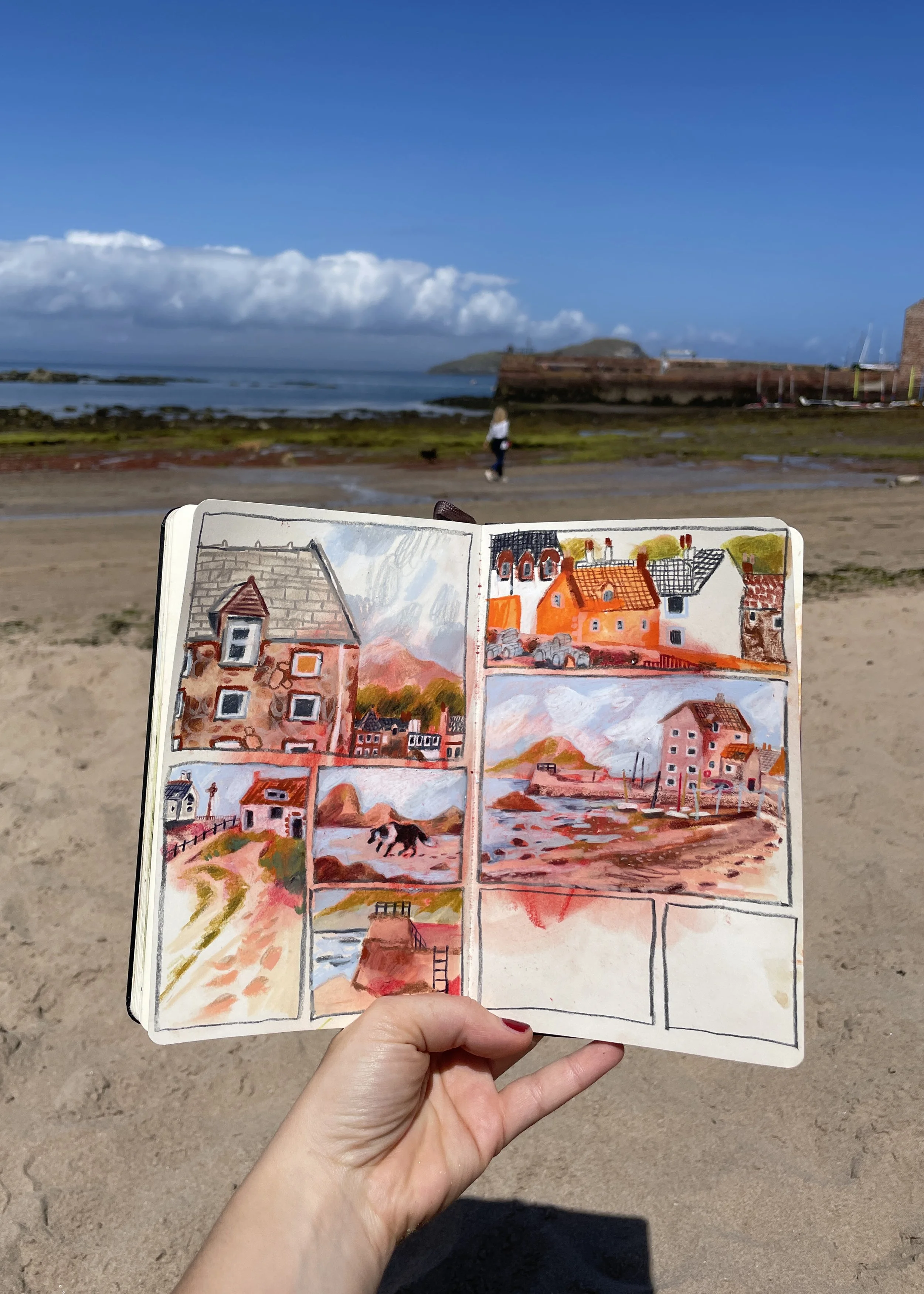 A person holding an open sketchbook with watercolor paintings of buildings and landscapes in front of a beach scene with blue sky, clouds, and a woman walking near rocks and a pier.