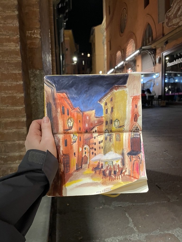 A sketch of a European-style street scene at night, with warmly lit buildings, outdoor seating, and a group of people sitting at a table under an umbrella.
