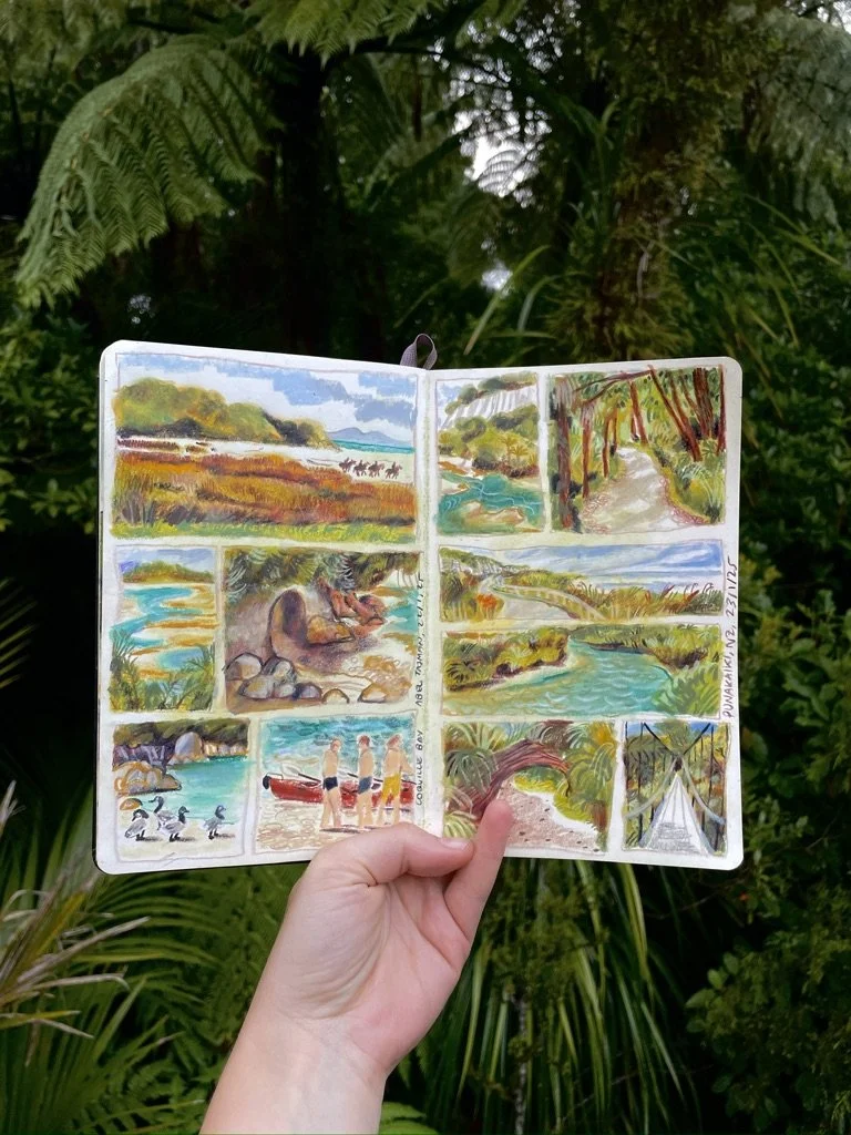 Hand holding a sketchbook with watercolor paintings of a river and nature scenes in a lush green outdoor setting.