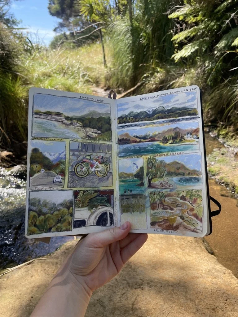 A hand holding an open sketchbook with watercolor sketches of nature scenes, including mountains, lakes, and a bicycle, against a natural outdoor background with trees, grass, and a small stream.