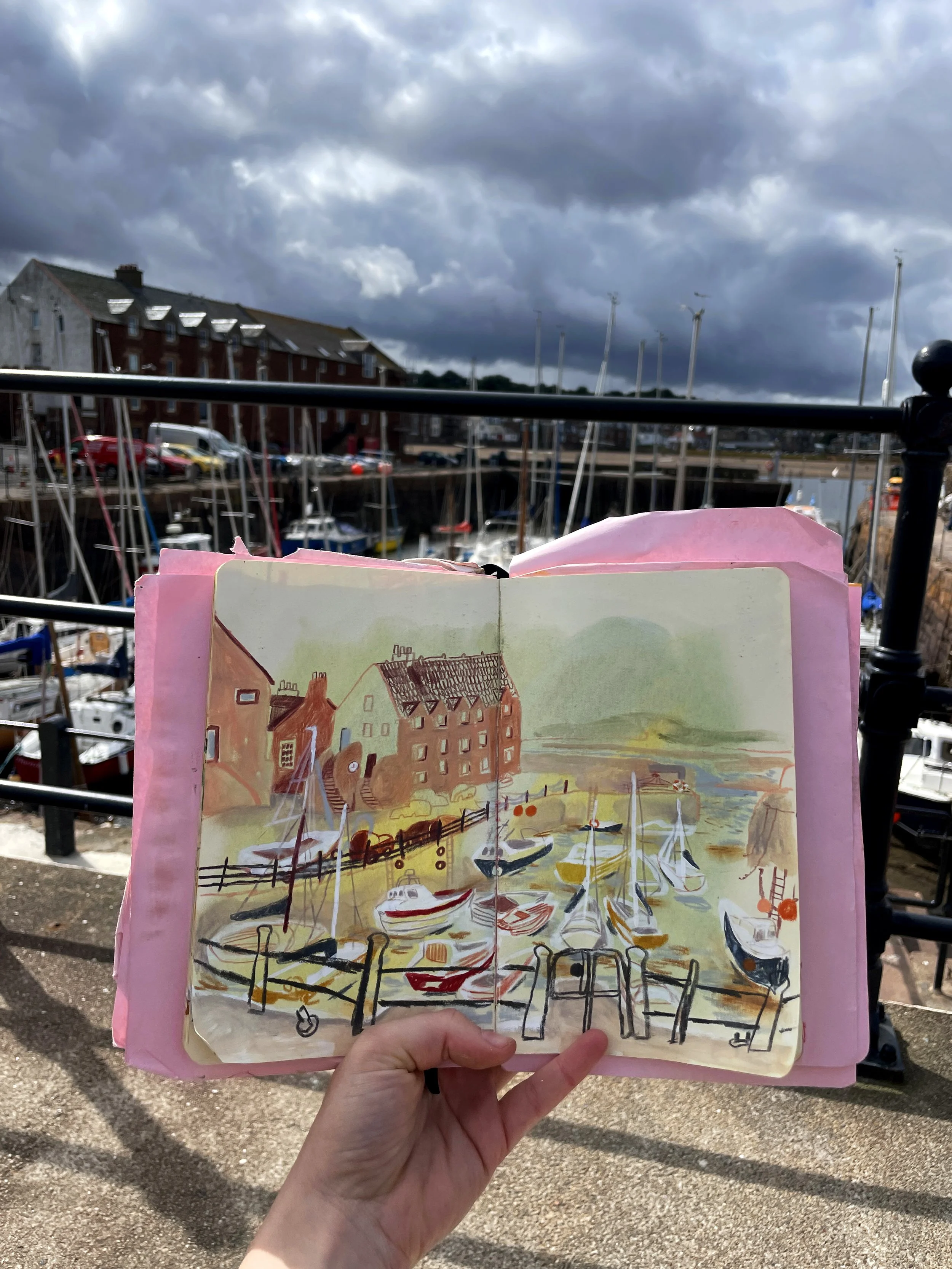 A person holding a watercolor painting of a marina with boats, houses, and fences, with a cloudy sky in the background.