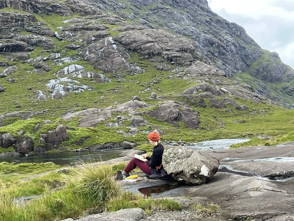 A woman sitting on a rock by a stream in a mountainous landscape, reading a book, surrounded by lush green grass and large rocks.