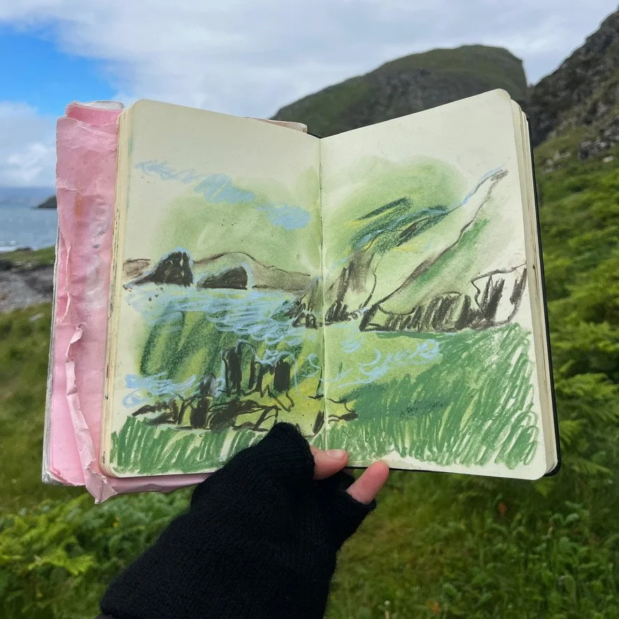 Sketch of a mountain landscape with green grass, rocks, and a body of water, held in front of a natural mountain view.