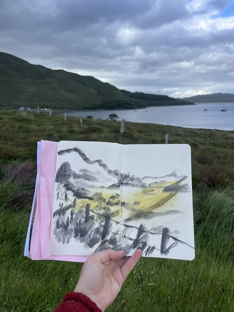 Hand holding a sketchbook with a landscape drawing of mountains and a fence, with a scenic lakeside view and mountains in the background under a cloudy sky.