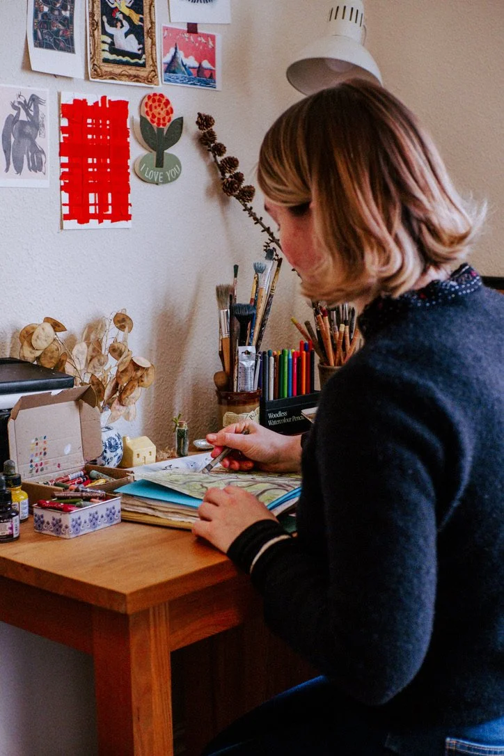 A person with shoulder-length hair sitting at a wooden desk, drawing in a sketchbook with various art supplies and paint bottles around. The wall behind them has art prints, a red paper craft, and decorative items.