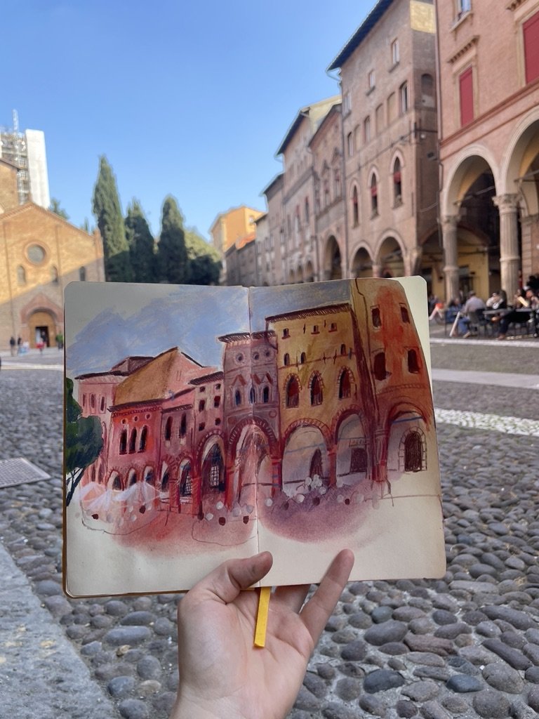 A person holds an open sketchbook with a watercolor painting of an old European building with arches, towers, and red walls, in front of the real building and a cobblestone street under a clear blue sky.