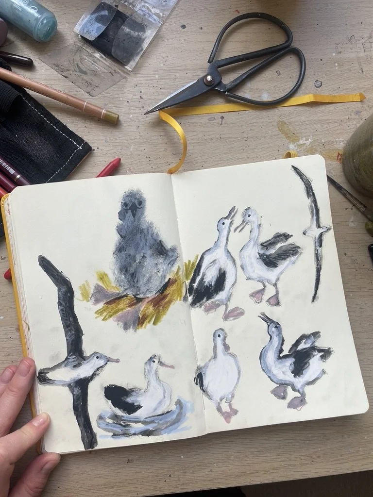 Open sketchbook with watercolor illustrations of ducks and a bird, surrounded by art supplies on a wooden table such as scissors, pens, and markers.