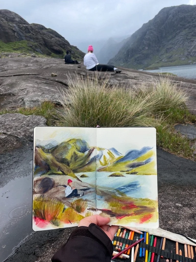 A sketchbook with a colorful landscape drawing of mountains, a river, and grassy terrain, held by a person outdoors. In the background, two people sit on rocks overlooking a mountainous lake scene.