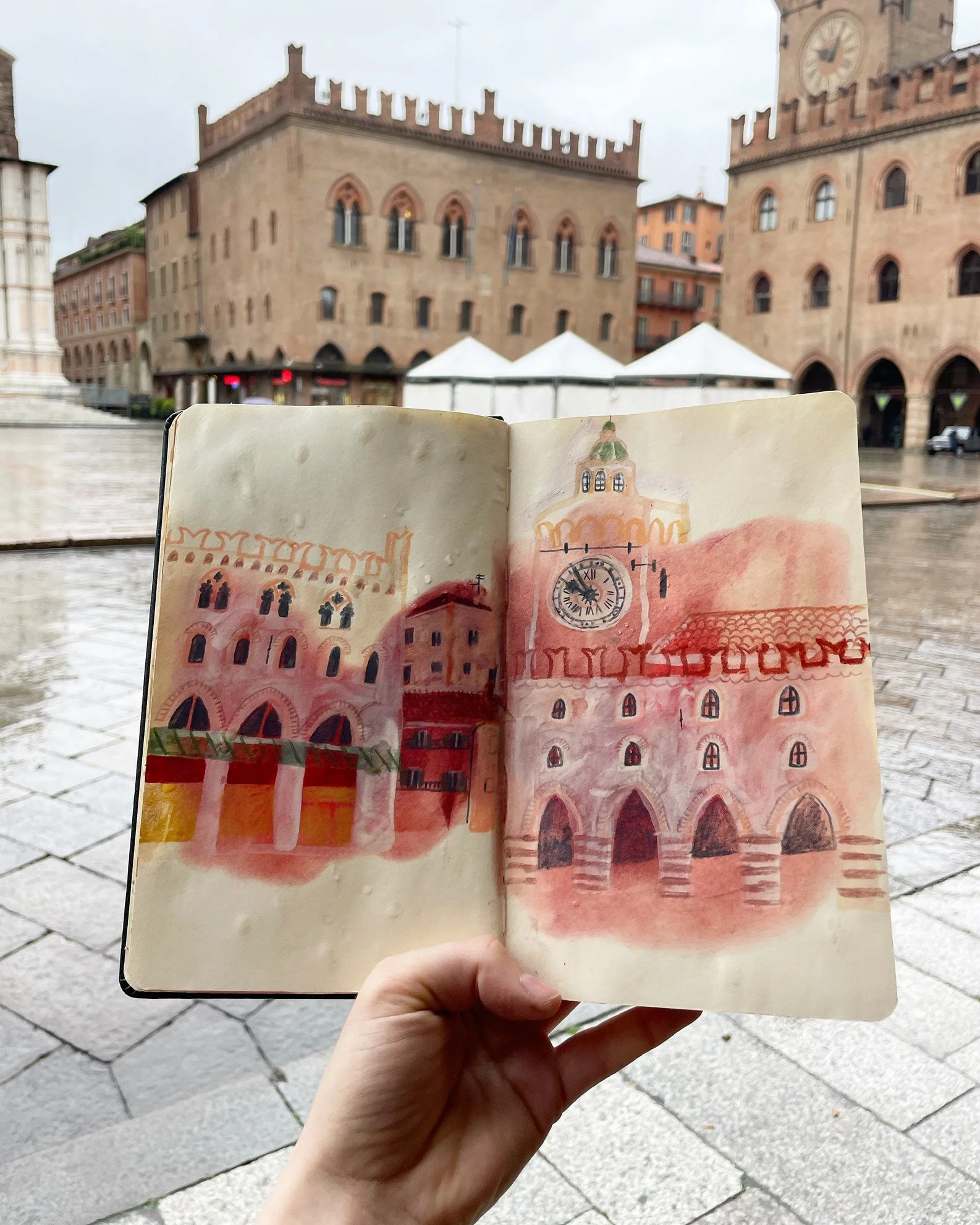 A hand holding a watercolor drawing of a historic building with arched windows and a clock, against a backdrop of old buildings, tents, and cloudy sky in an urban square.