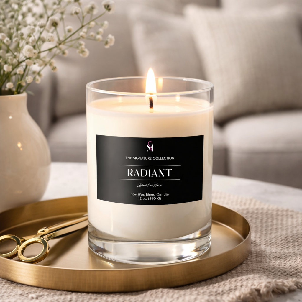 Lit candle labeled 'Radiant' on a gold tray, with a pair of gold scissors beside it, on a beige woven tablecloth, with a white vase of white flowers and a gray sofa in the background.