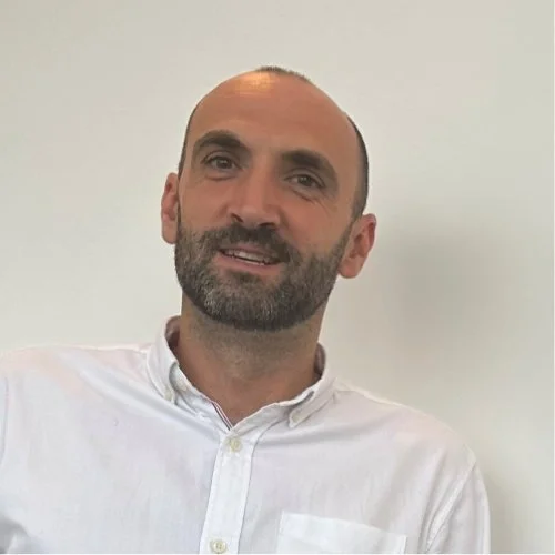 A man with a beard and short hair wearing a white dress shirt, smiling, against a plain light-colored wall.