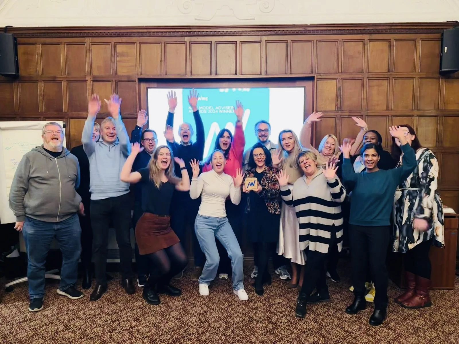Group of people celebrating with raised hands, smiling, in a conference room with a wooden paneled wall and a large digital screen behind them.
