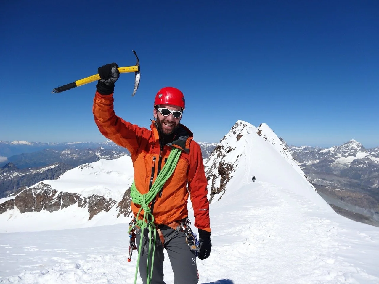 A climber in orange jacket, red helmet, and sunglasses holding an ice axe standing on snowy mountain summit with snow-capped peaks in the background.