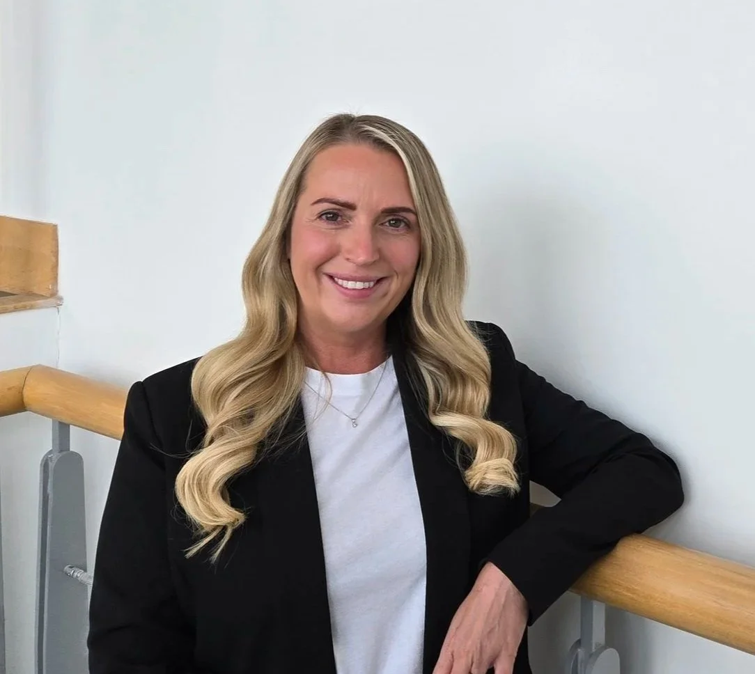 A woman with long blonde hair wearing a black blazer and white shirt, smiling, leaning against a wooden handrail.