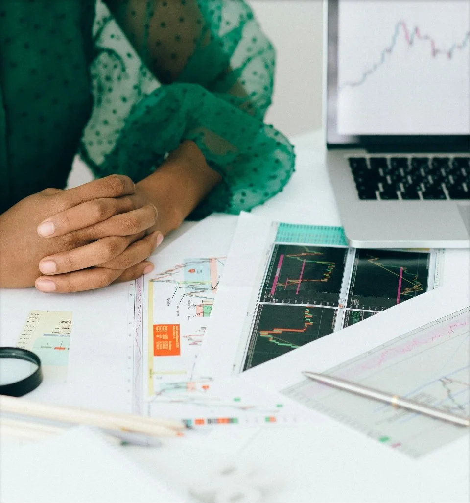 Person sitting at desk with financial charts, graphs, and a laptop showing stock market data.