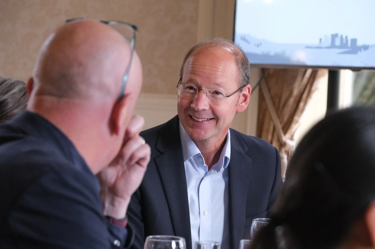 A man in glasses and a dark suit smiling while speaking with others at a dinner or meeting.