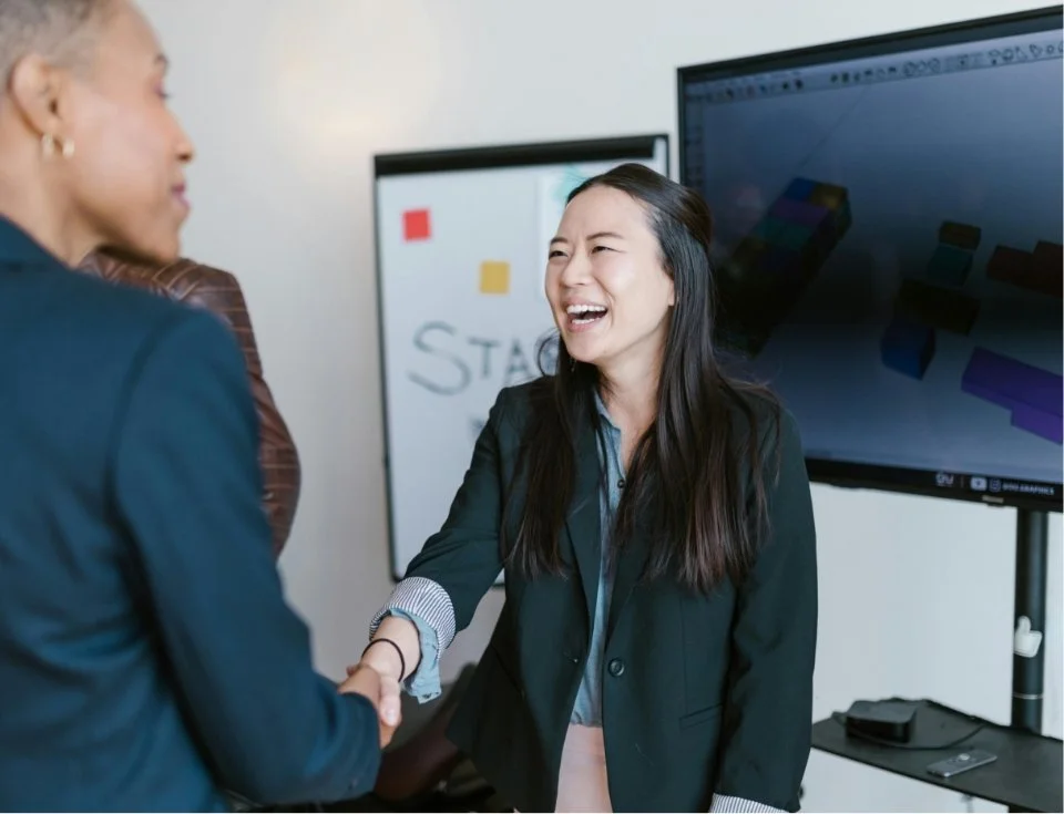 Two women shaking hands and smiling in a business setting, with a large monitor and a whiteboard in the background.
