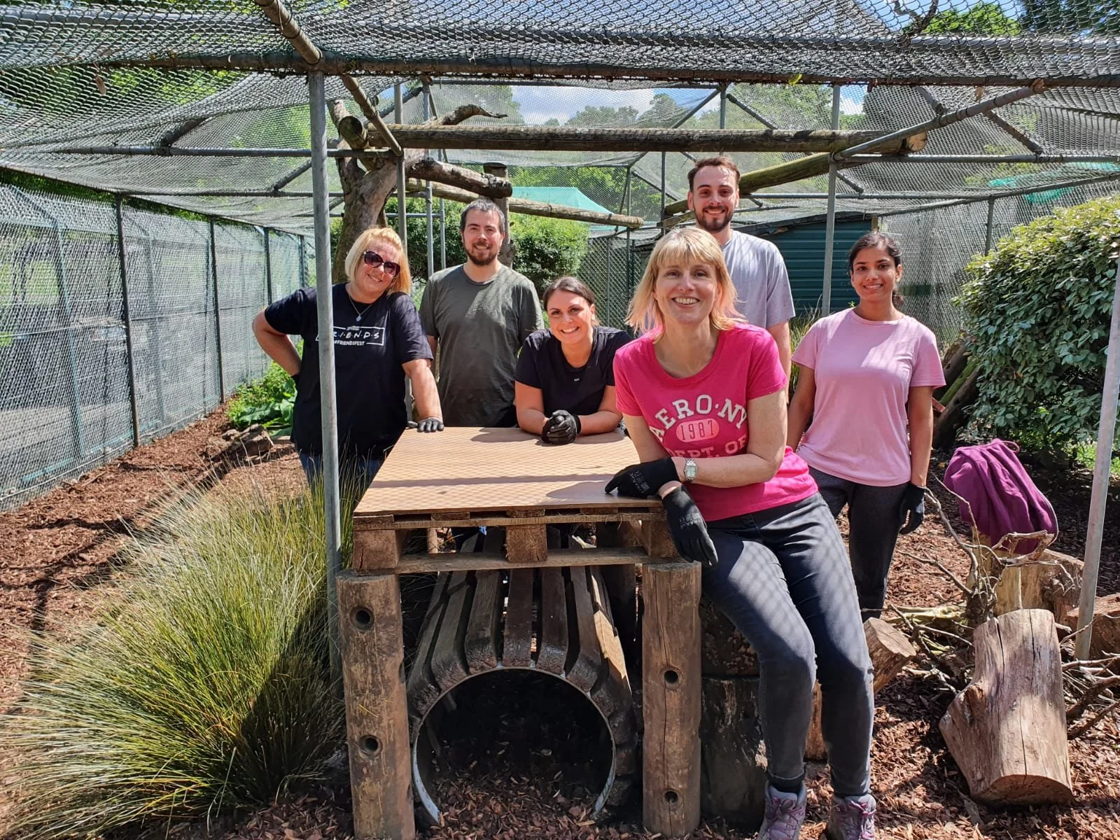 Group of six people working in a garden or outdoor project, standing and sitting around a wooden table, with a shade cover overhead, plants and green bushes in the background, smiling and wearing work gloves.