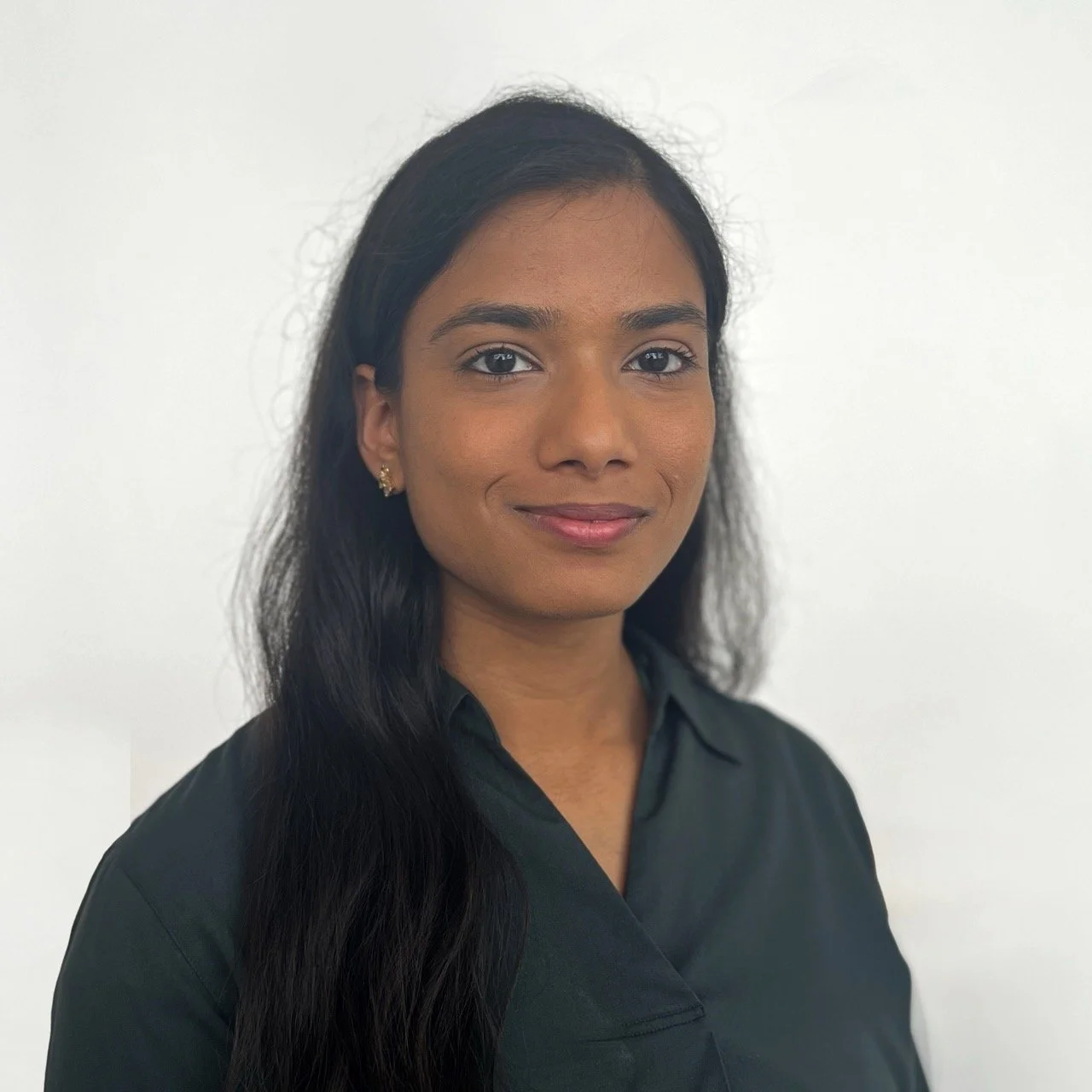 A young woman with long black hair, wearing a black shirt and small earrings, smiling against a plain white background.