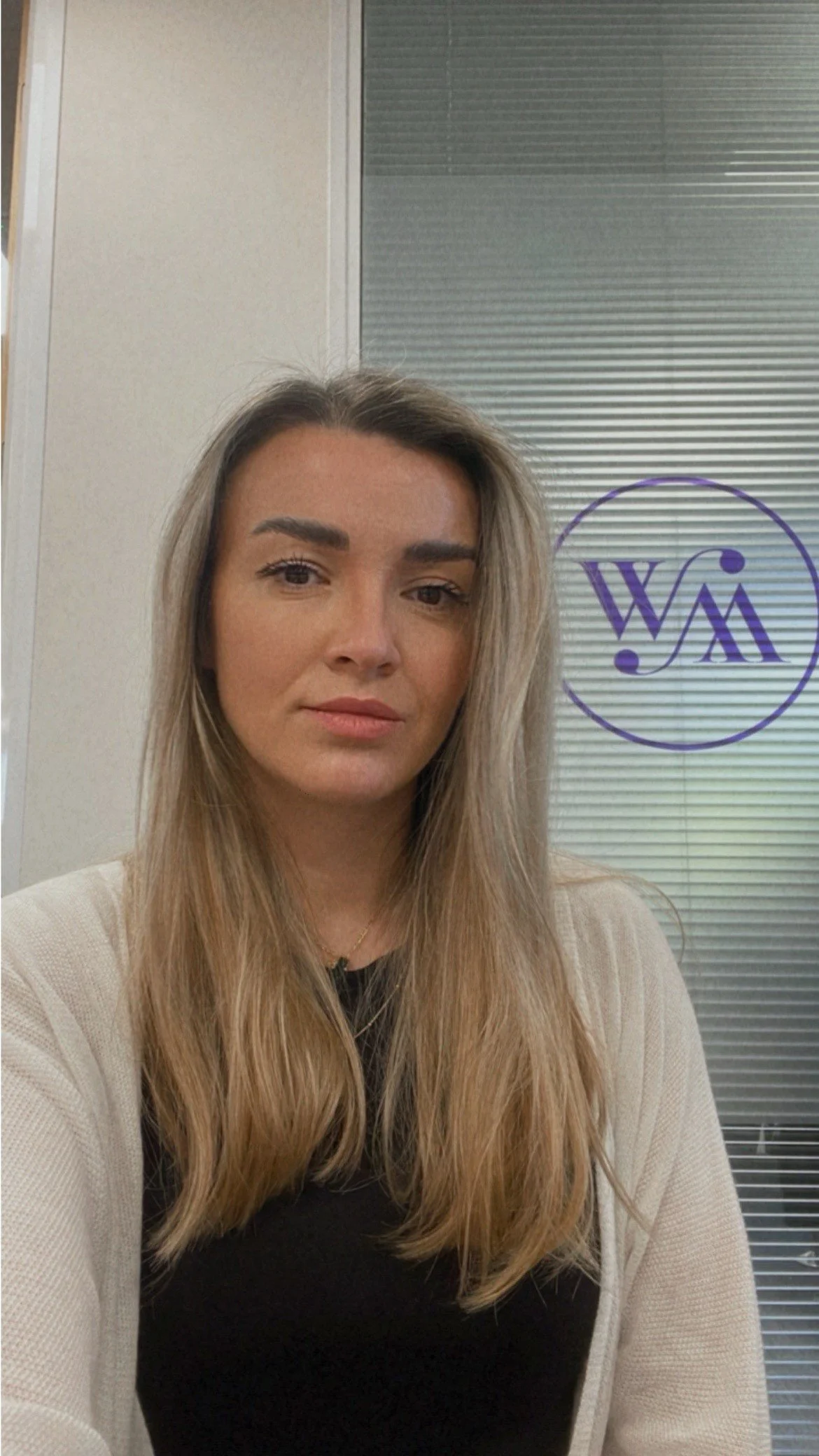 A woman with long, light brown hair wearing a black top and beige cardigan, taking a selfie in an office with a gray wall and a window with blinds behind her.