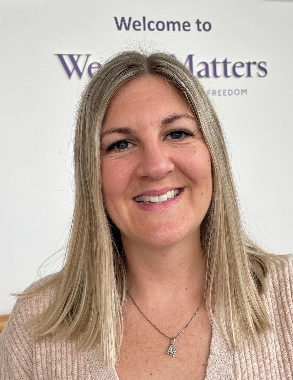 A smiling woman with blonde hair wearing a light pink sweater and a letter "M" necklace, standing in front of a sign that says "Welcome to We Matter".