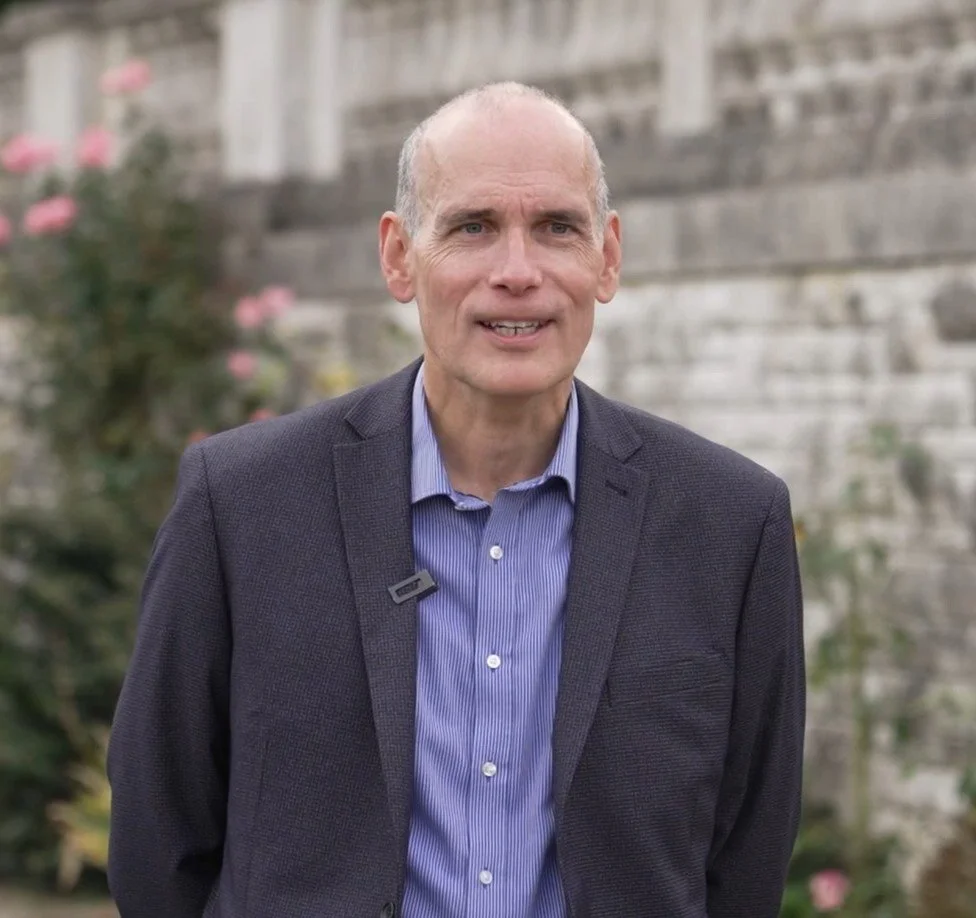 Man in a suit standing outdoors in front of a stone wall and pink flowers.