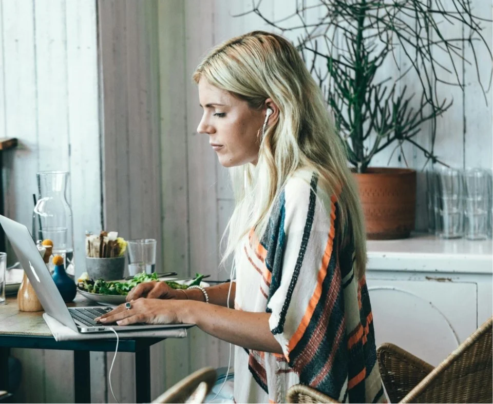 Woman working on laptop at a desk with a salad and a water pitcher in a cozy, wooden-themed room.