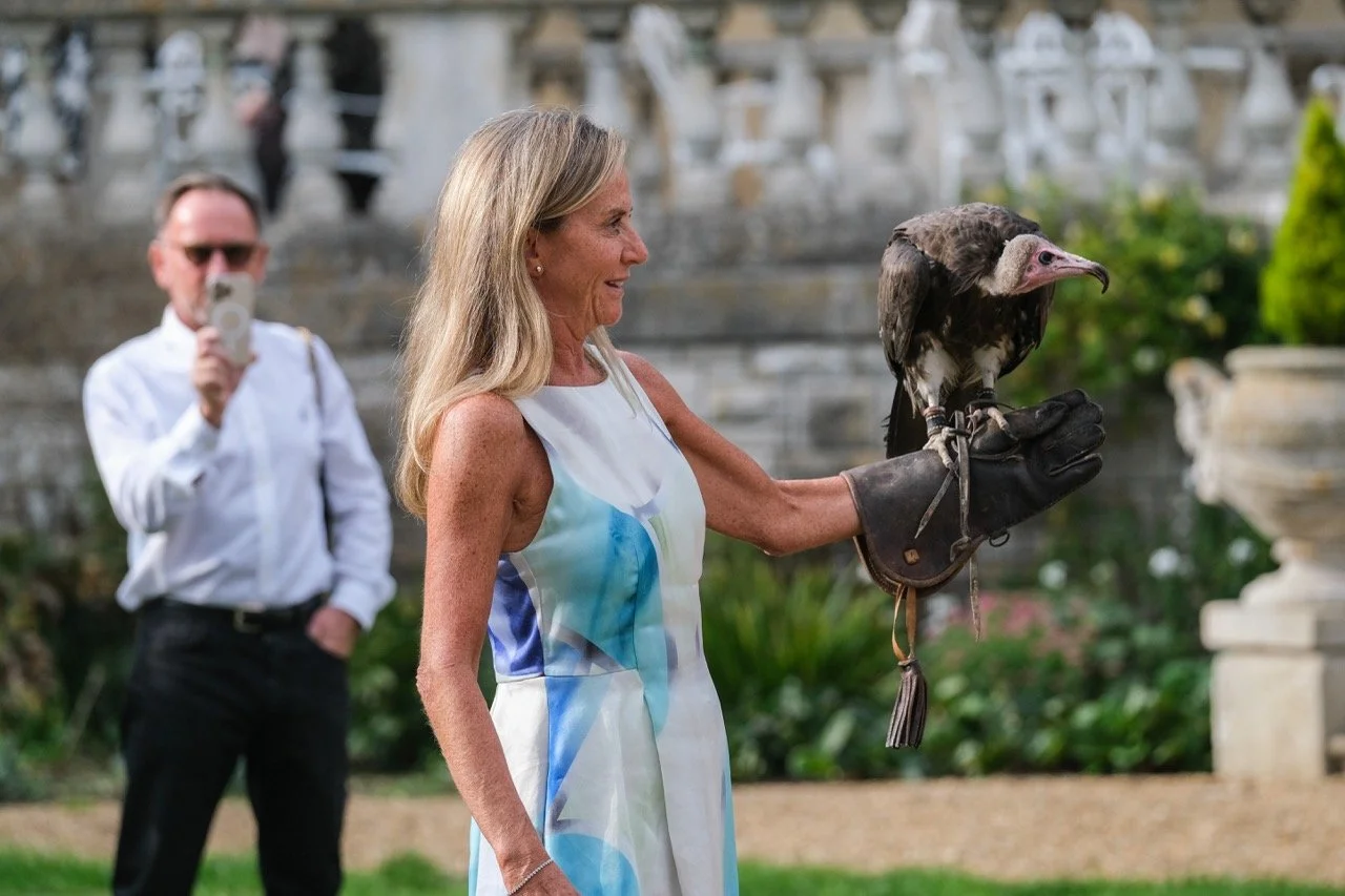 A woman holding a vulture on her gloved hand while a man in the background records with a cell phone at an outdoor location with stone structures and greenery.
