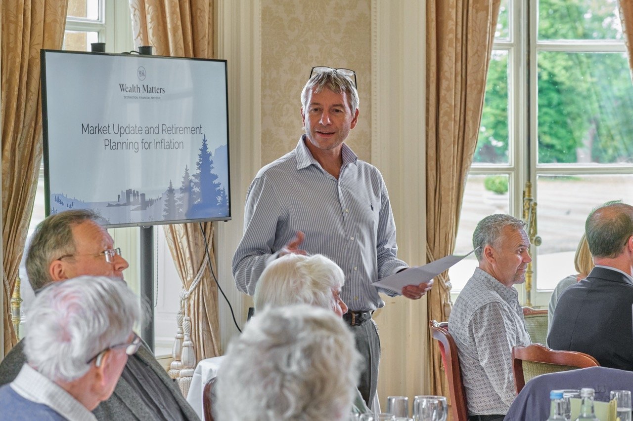 A man presenting at a conference, standing by a screen displaying a slide titled 'Market Update and Retirement Planning for Inflation,' with audience members seated at tables.