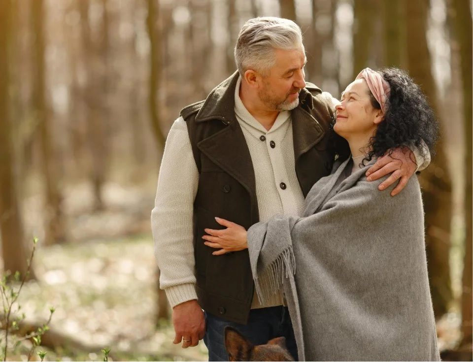 An elderly man and woman embrace and look into each other's eyes in a forest during autumn.