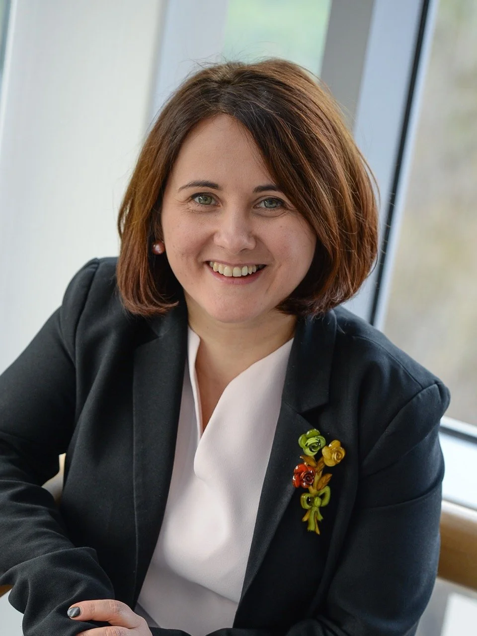 A woman with shoulder-length reddish-brown hair smiling, wearing a black blazer with colorful floral pins, sitting at a table near a window in an office setting.