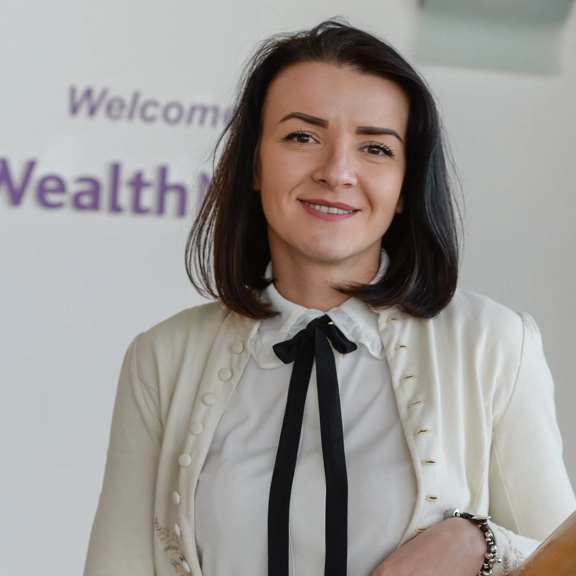 A woman with dark hair smiling, wearing a white blouse and jacket with a black bow tie, standing in front of a sign that says 'Welcome Wealth'.