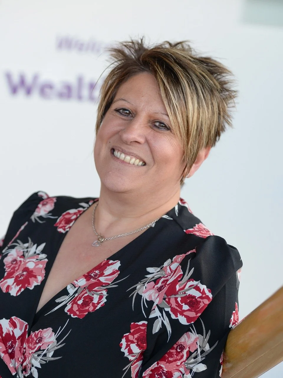 Smiling woman with short blonde hair, wearing a black dress with pink floral pattern and a silver necklace, standing indoors.