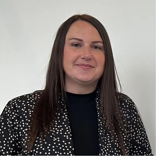 A woman with long brown hair, wearing a black top and a black and white polka dot jacket, standing against a plain white background.