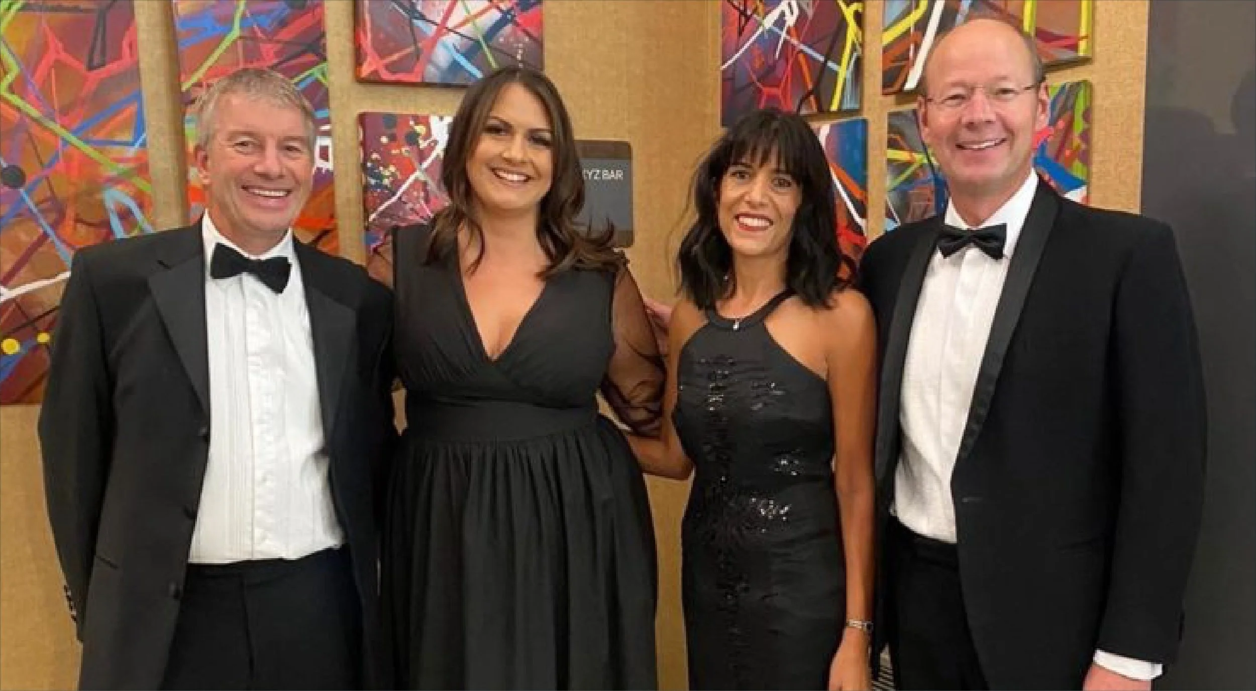 Four people dressed in formal attire, two men in tuxedos and two women in elegant black dresses, standing together and smiling at an art gallery with colorful abstract paintings in the background.
