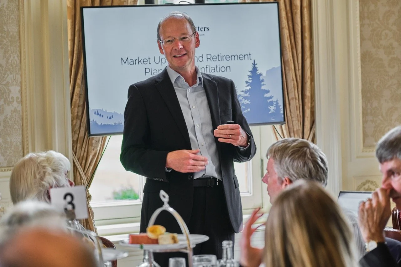 Man in a dark suit giving a presentation at a conference or event with a large screen behind him displaying the words 'Market Up and Retirement Planning.' Audience members are seated at tables with food and drinks.