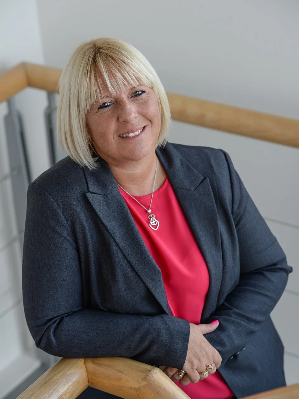 A middle-aged woman with blonde hair and a short bob haircut, smiling, wearing a dark gray blazer over a pink blouse with a silver heart-shaped necklace, standing by a wooden handrail in a modern interior.