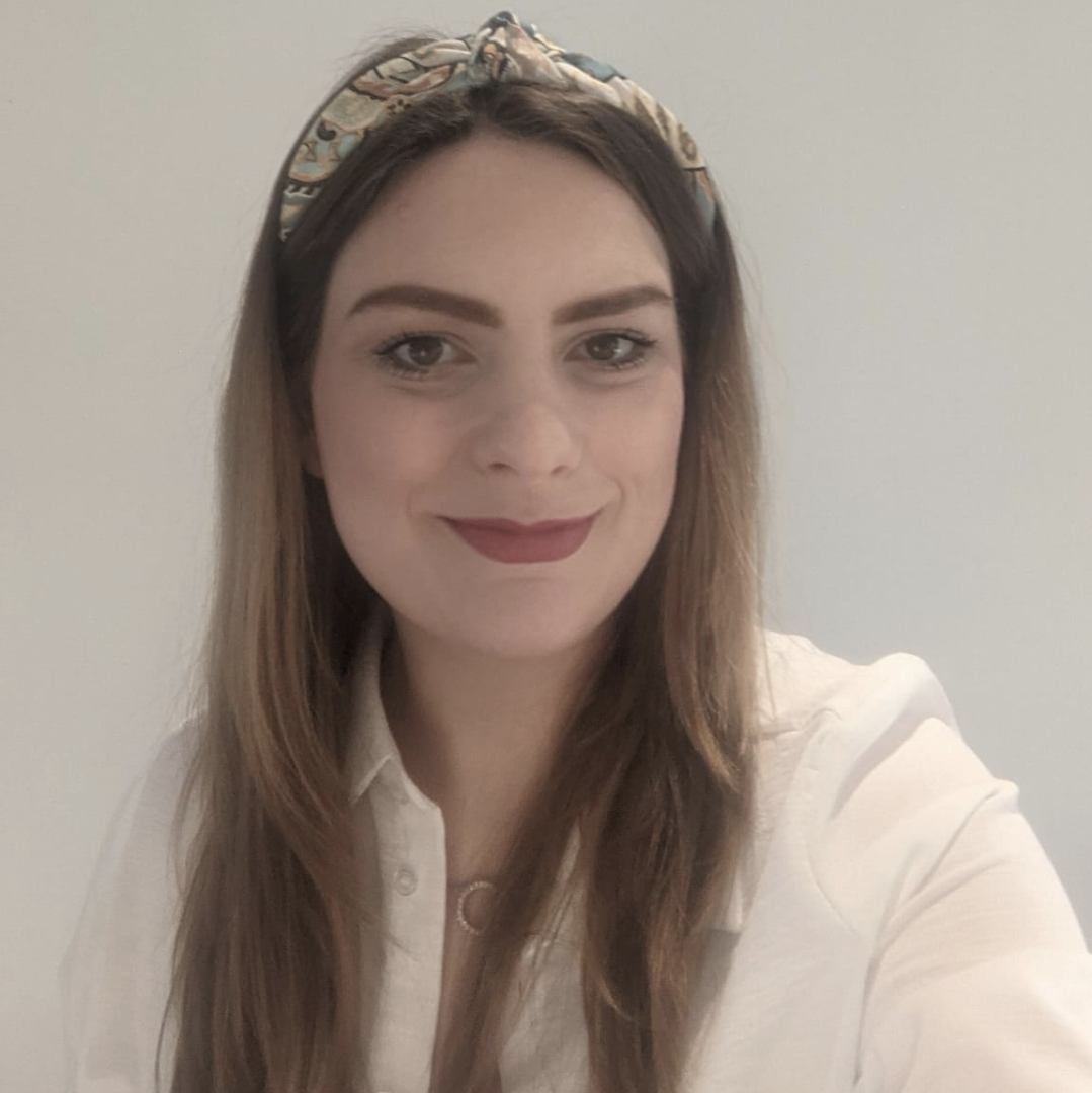 A young woman with long brown hair, wearing a patterned headband and a white button-up shirt, smiling in front of a plain light-colored wall.