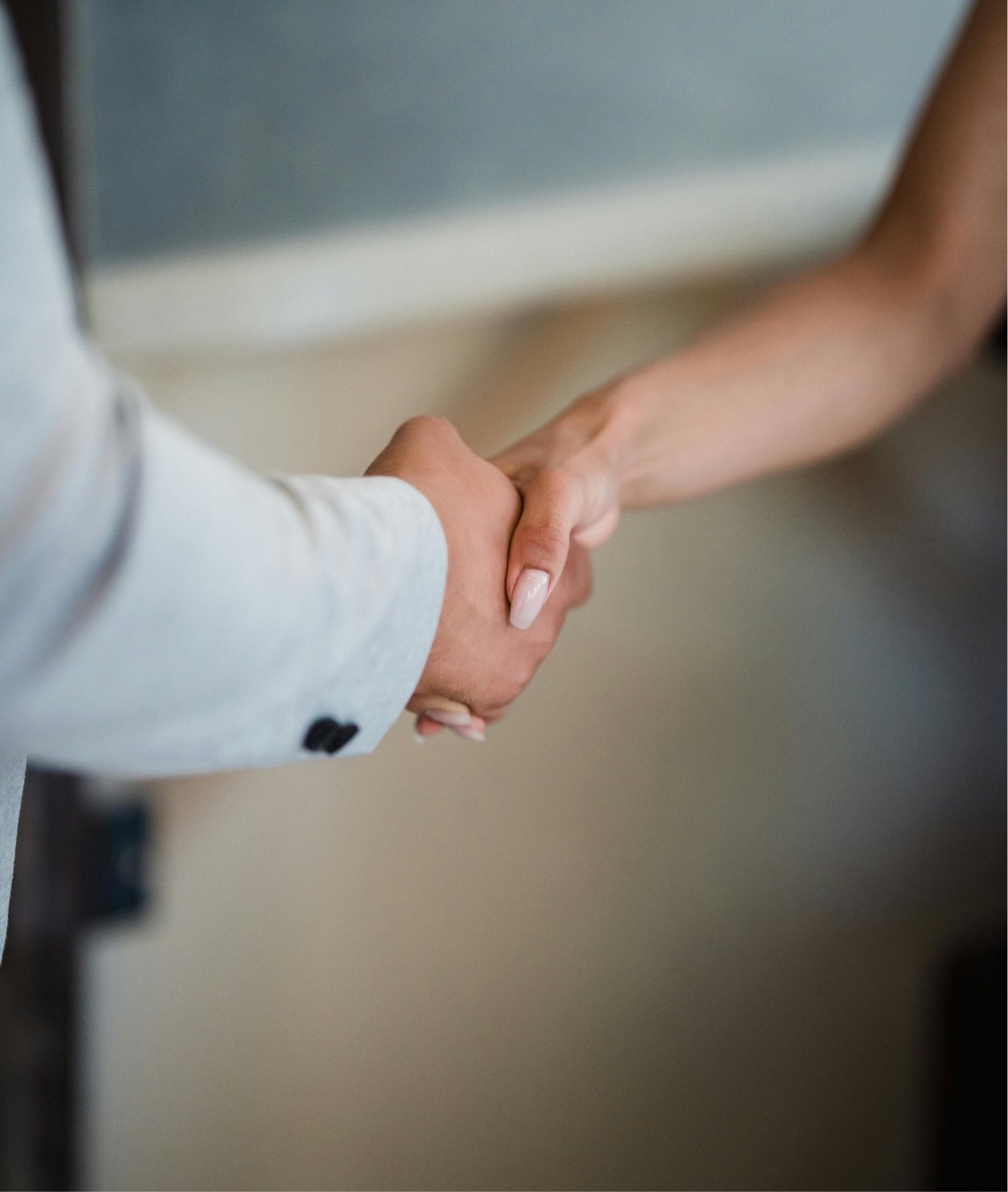Close-up of two people shaking hands, one wearing a light-colored suit jacket with a buttoned cuff, the other with a bare arm, indoors.