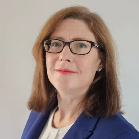 A woman with shoulder-length light brown hair, wearing glasses, a white top, and a blue blazer, smiling softly against a plain light background.