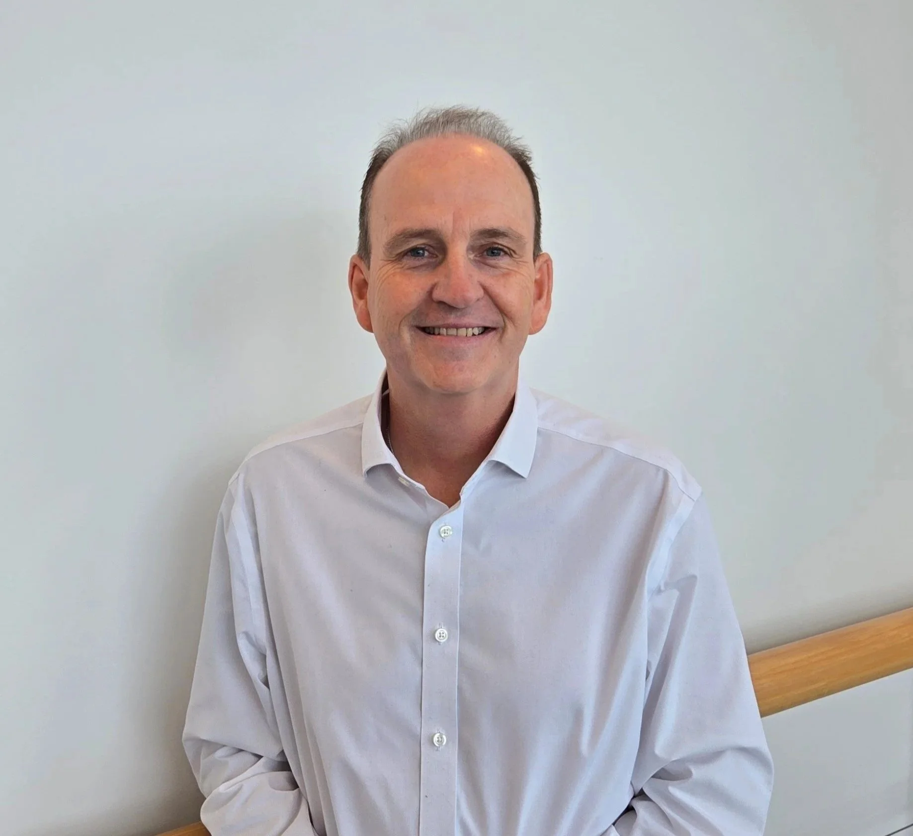 A smiling man with short, receding hair wearing a white button-up shirt, standing against a plain white wall with a wooden handrail beside him.