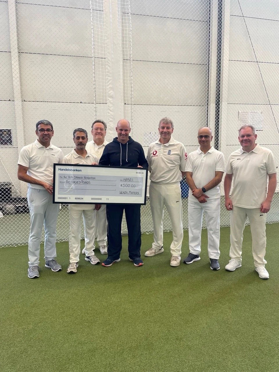 Group of seven men standing on an indoor cricket pitch, holding a large check for 500 pounds made out to The Run Strauss Foundation, celebrating a donation.